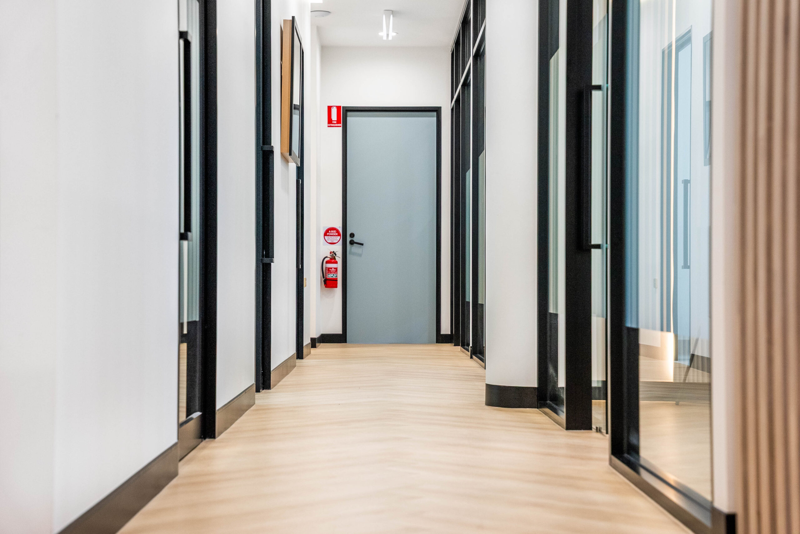 Wide clinic corridor with level flooring, glazed treatment room doors and clearly visible fire safety equipment